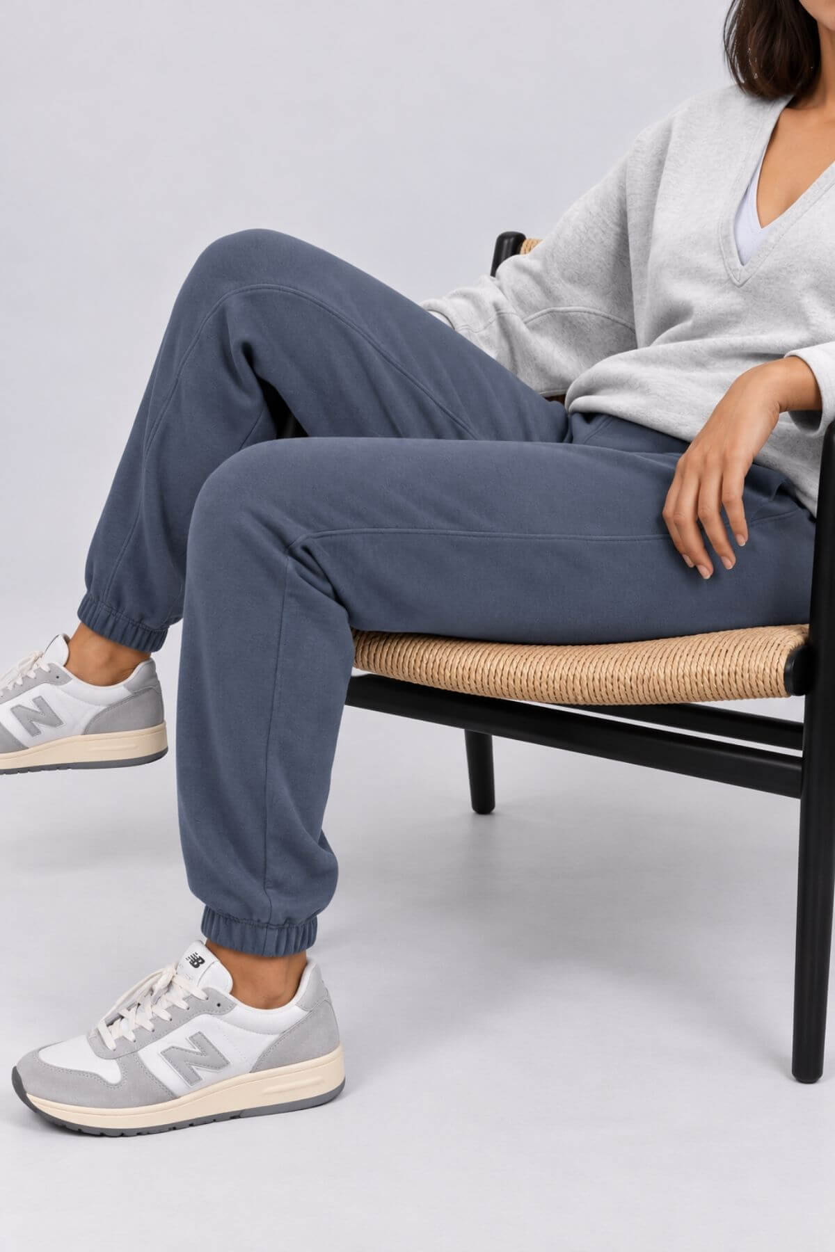 A person seated on a chair wearing slate blue jogger sweatpants, a light gray sweater, and gray New Balance sneakers against a light gray studio background.