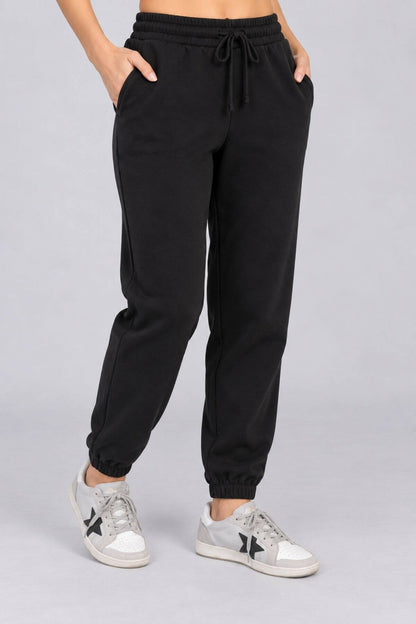 Black jogger sweatpants with a drawstring waist and elastic cuffs, shown on a model wearing light gray sneakers against a light gray studio background.