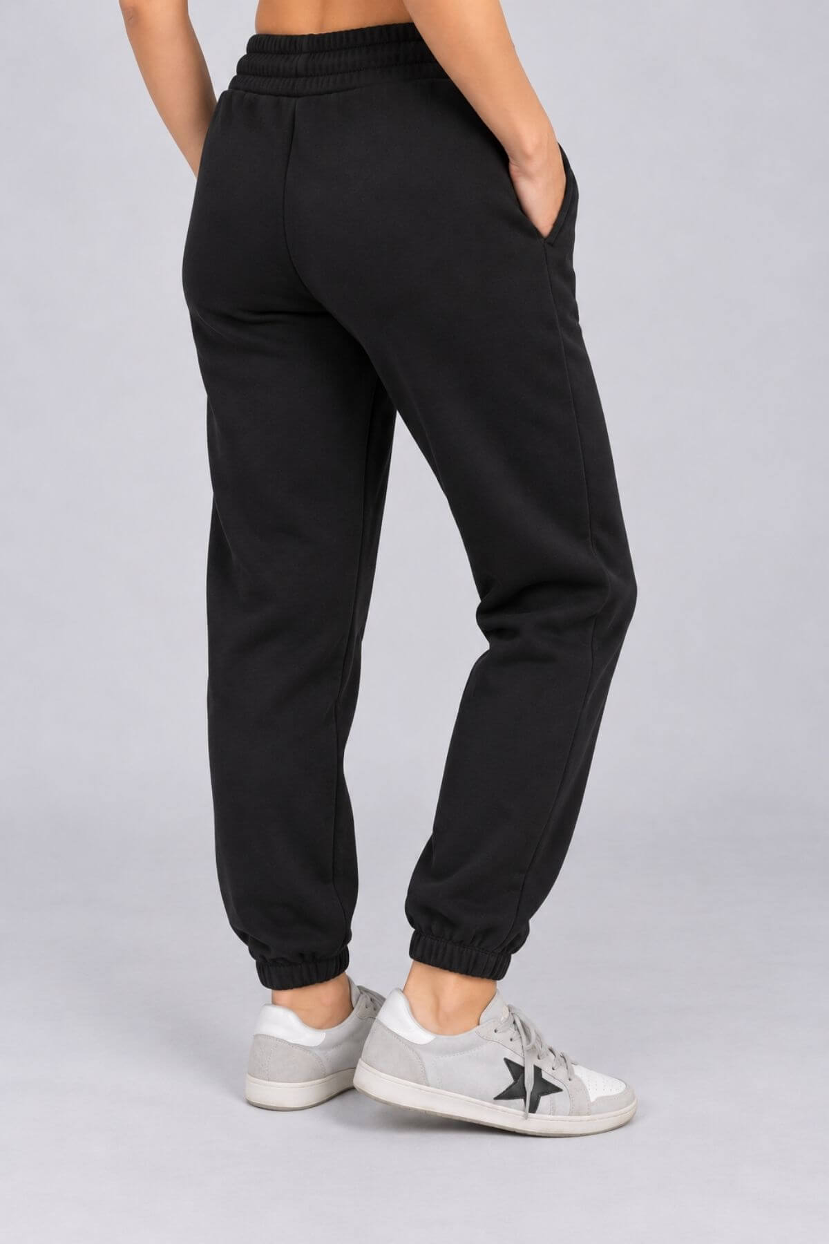 Black jogger sweatpants worn by a model, shown from the back three-quarter view against a light gray studio background. The model is wearing white and gray sneakers.