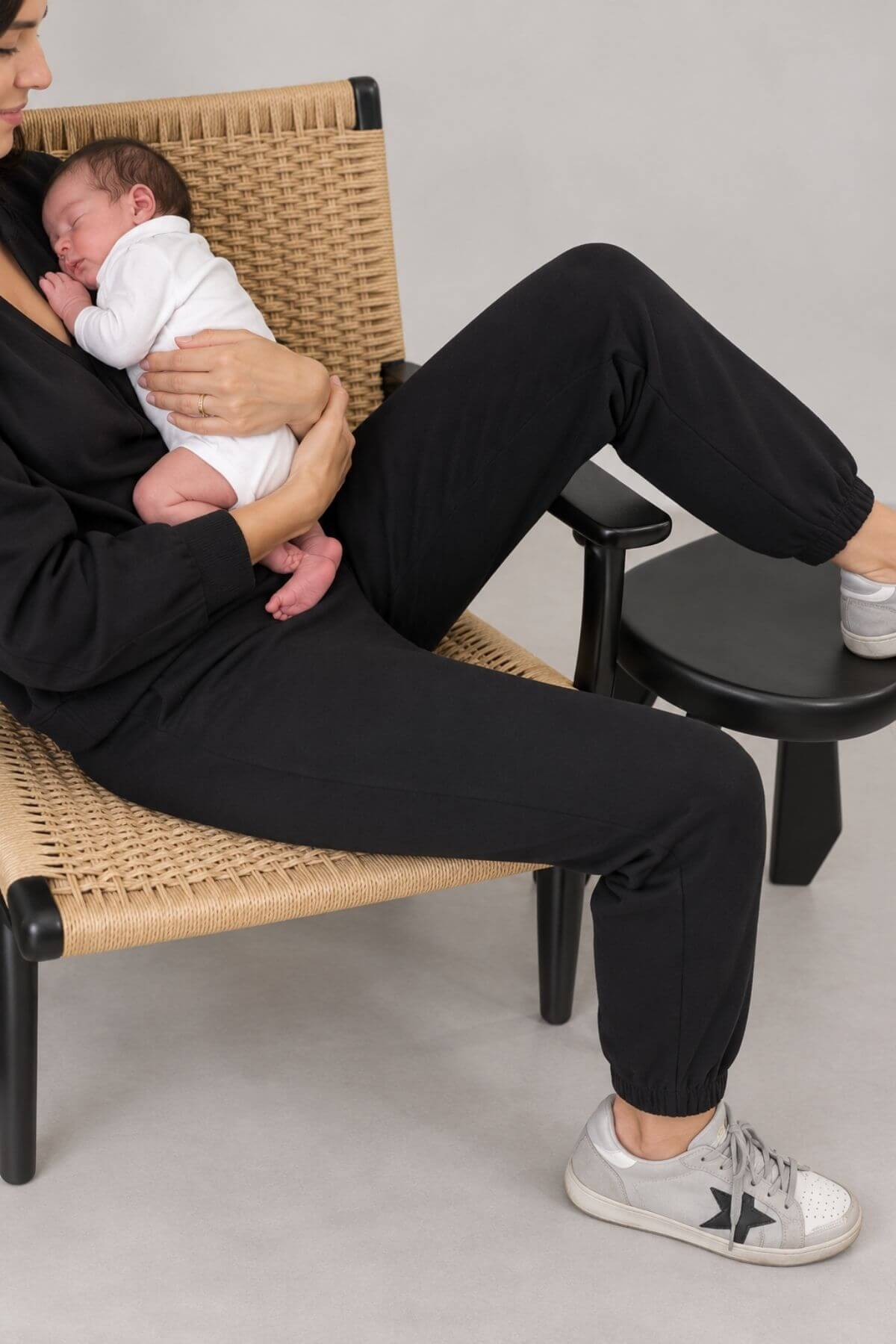 A person wearing a black loungewear set with jogger pants and white sneakers sits on a woven lounge chair while holding a sleeping newborn baby in a white swaddle. The scene is set against a neutral gray studio background.