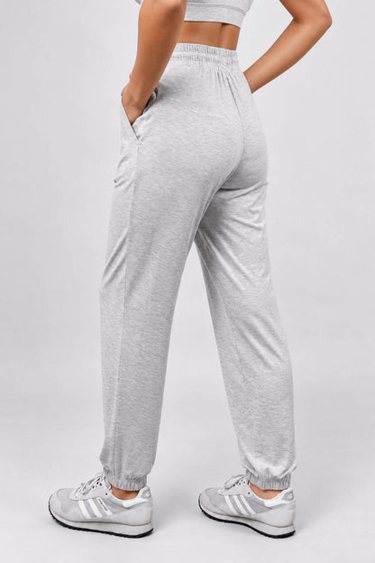A person wearing light heather gray jogger sweatpants with elastic cuffs and side pockets, paired with white sneakers, shown from the back against a light gray studio background.