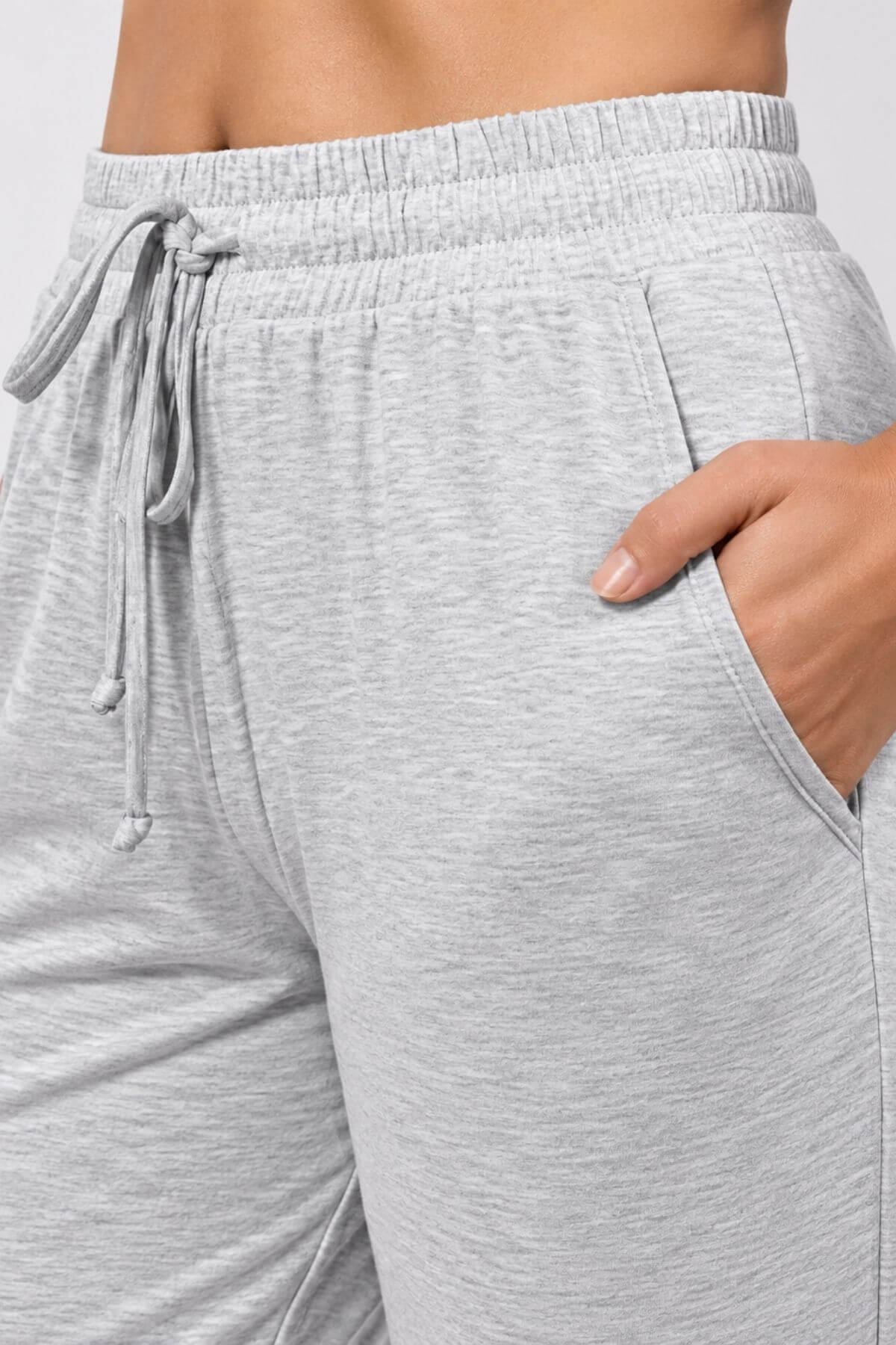 Close-up of a person wearing light heather gray drawstring sweatpants, showing the waistband, side pocket with a hand in the pocket, and part of the hip.