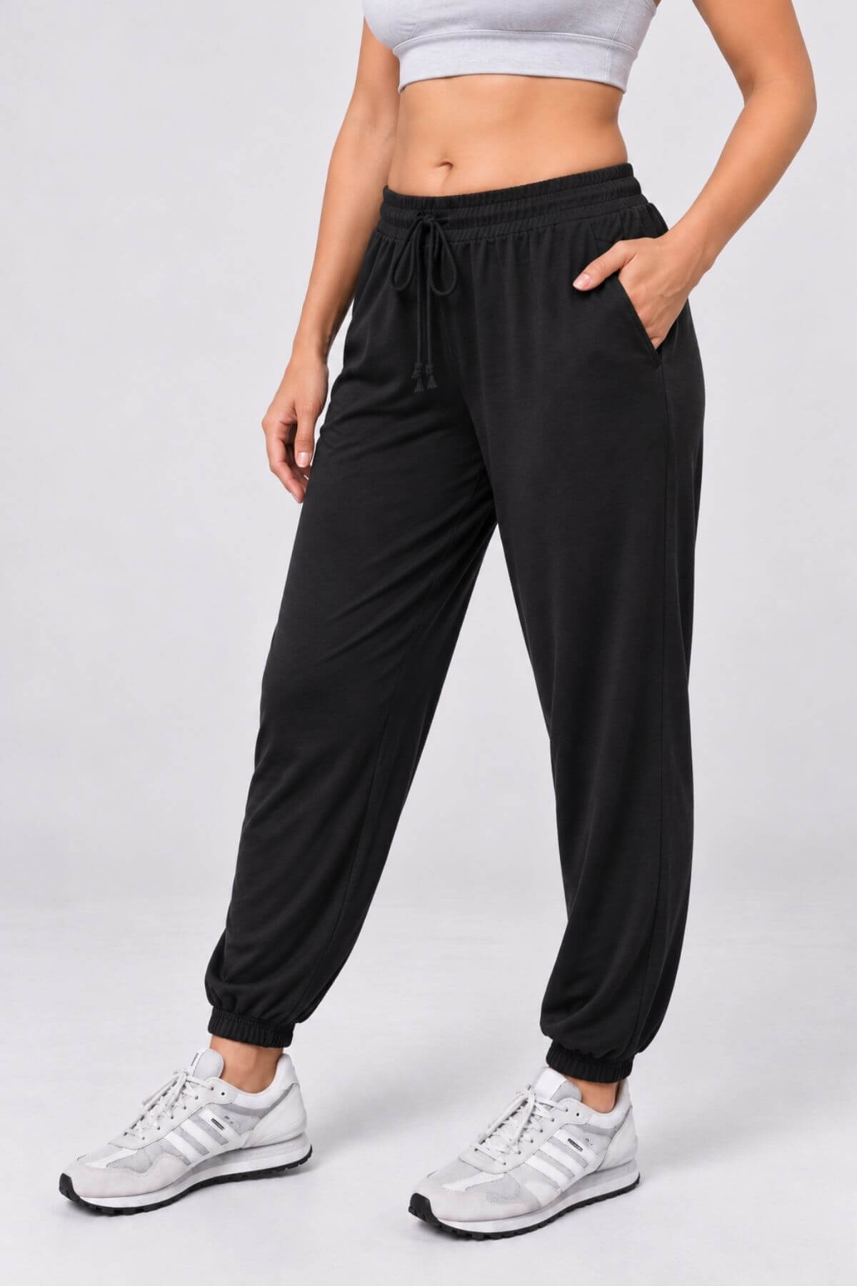 A model wearing black jogger sweatpants with a drawstring waist, side pockets, and elastic cuffs, paired with white sneakers, shown from the waist down against a light gray studio background.