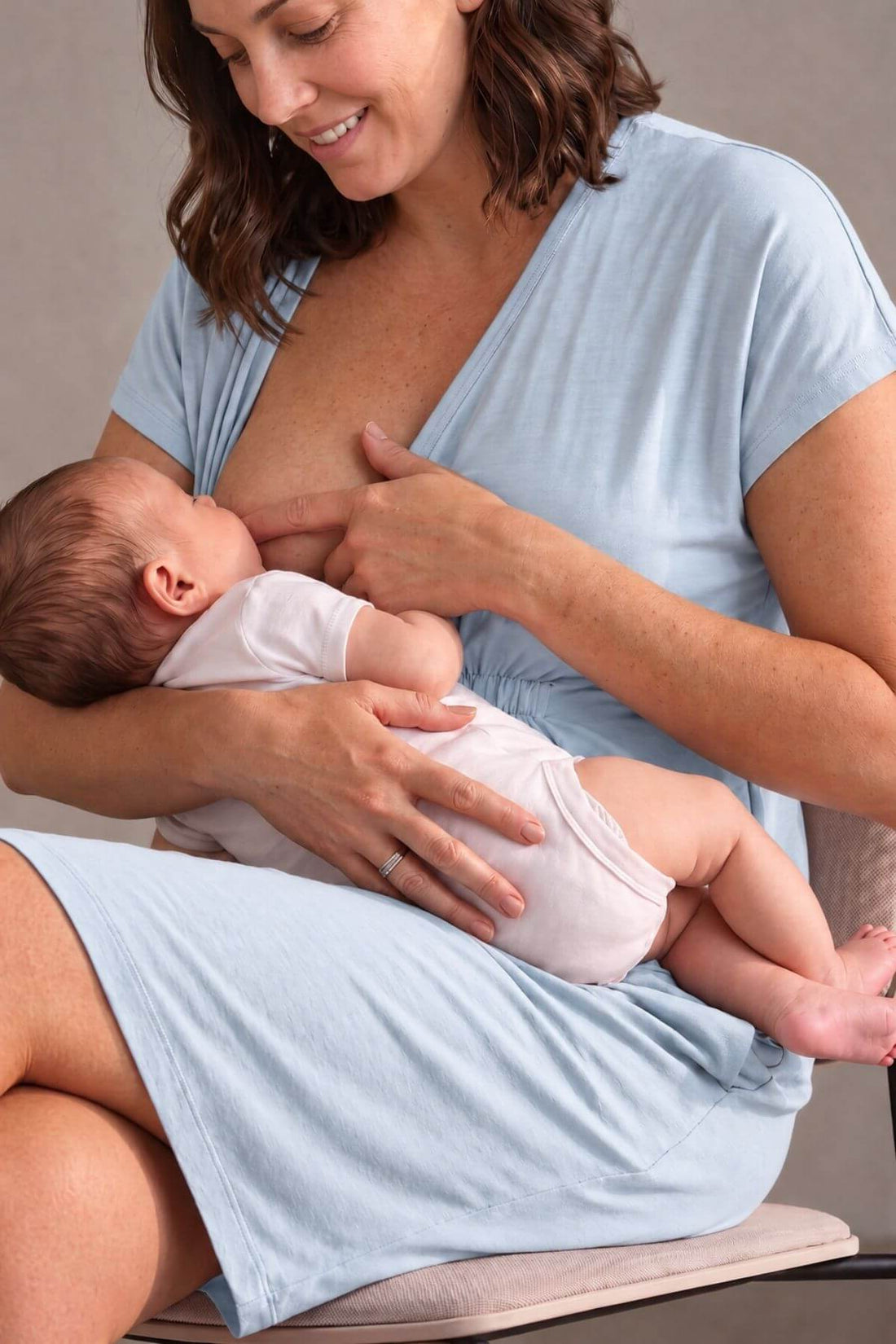 A woman in a light blue maternity dress breastfeeding a baby wearing a white onesie, seated on a chair in a softly lit indoor setting.