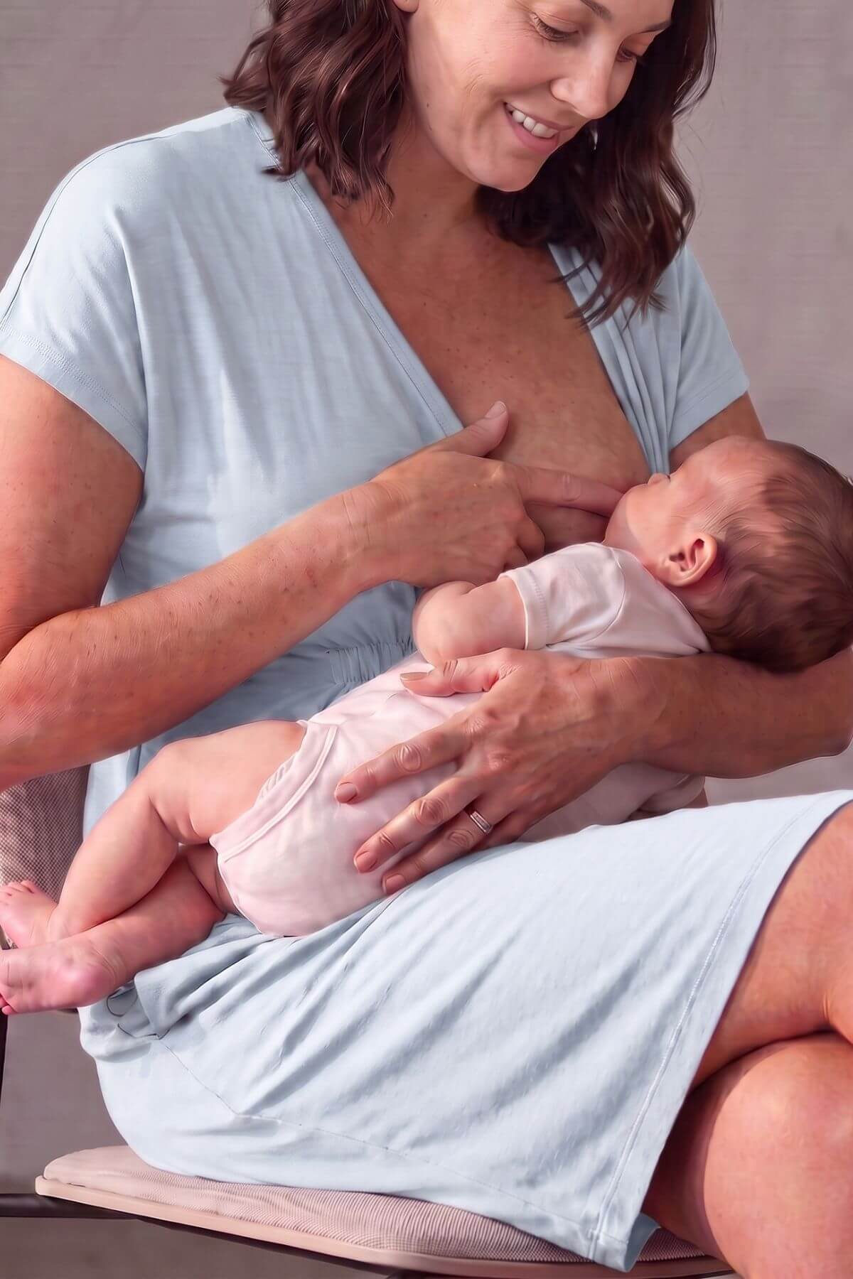 A woman in a light blue nursing dress holds a baby wearing a pale pink bodysuit while breastfeeding. The woman is smiling and looking down at the baby, and they are seated on a cushioned surface against a soft neutral background.