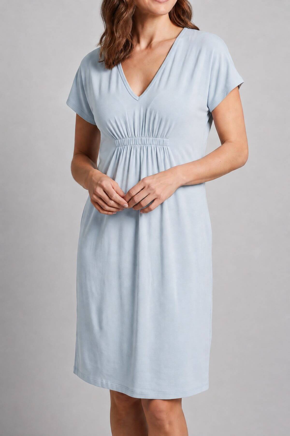 A woman wearing a light blue short-sleeve V-neck maternity dress with a gathered elastic waist, shown against a neutral gray studio background.