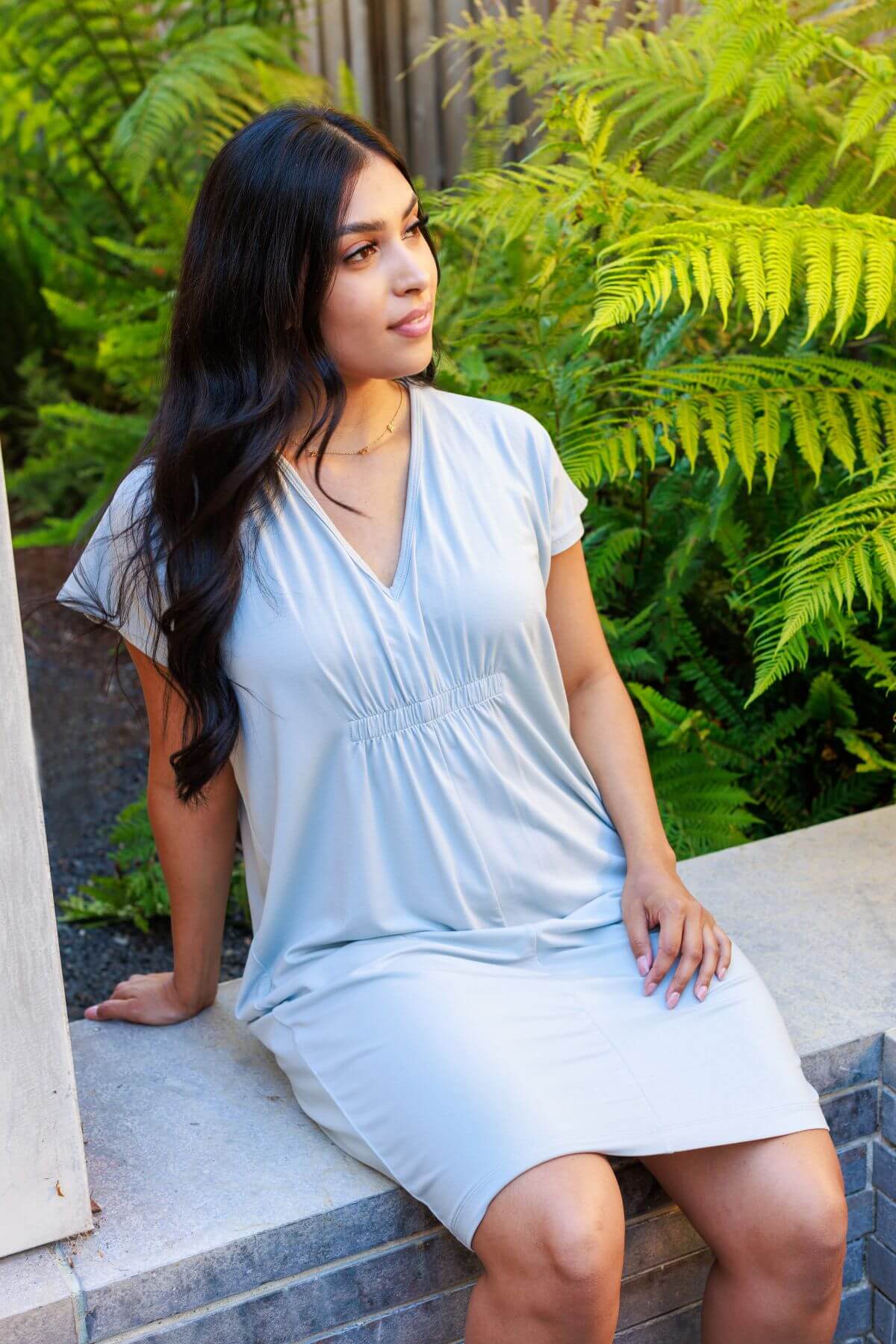 Woman in a light blue dress sitting on a stone ledge with greenery in the background