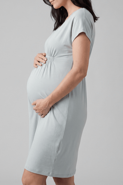 A pregnant woman wearing a light blue-gray short-sleeve maternity dress, shown from the side against a neutral gray studio background.