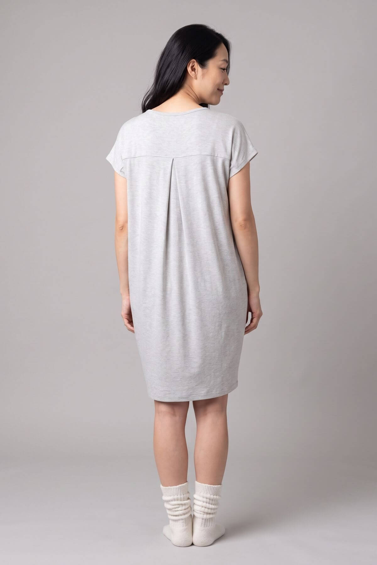 A woman viewed from the back wearing a light gray short-sleeve t-shirt dress, paired with white socks, against a solid light gray studio background.