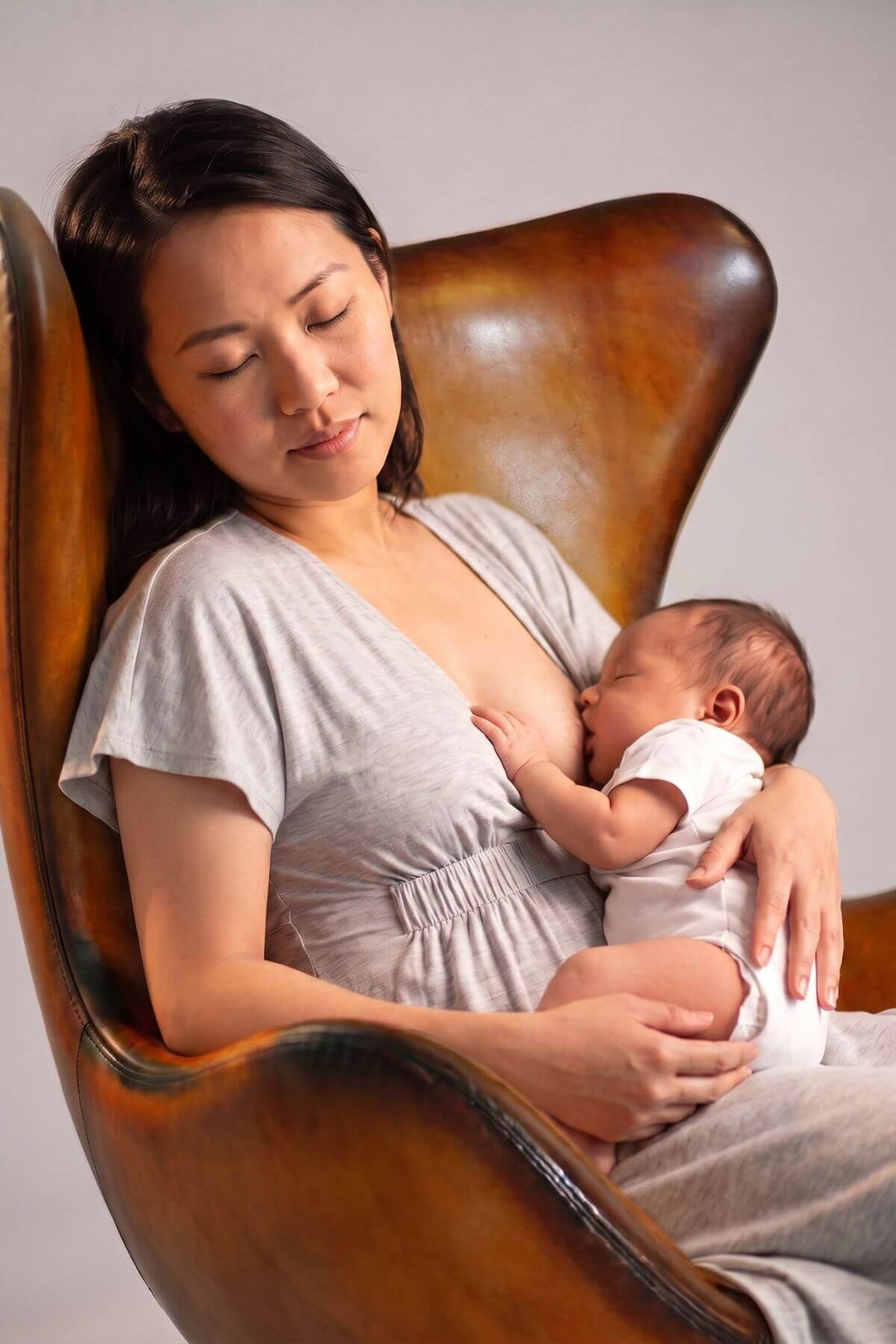 A woman in a light gray dress breastfeeding a baby wearing a white onesie, seated in a brown leather armchair against a plain light gray background.
