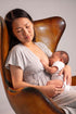 A woman in a light gray dress breastfeeding a baby wearing a white onesie, seated in a brown leather armchair against a plain light gray background.