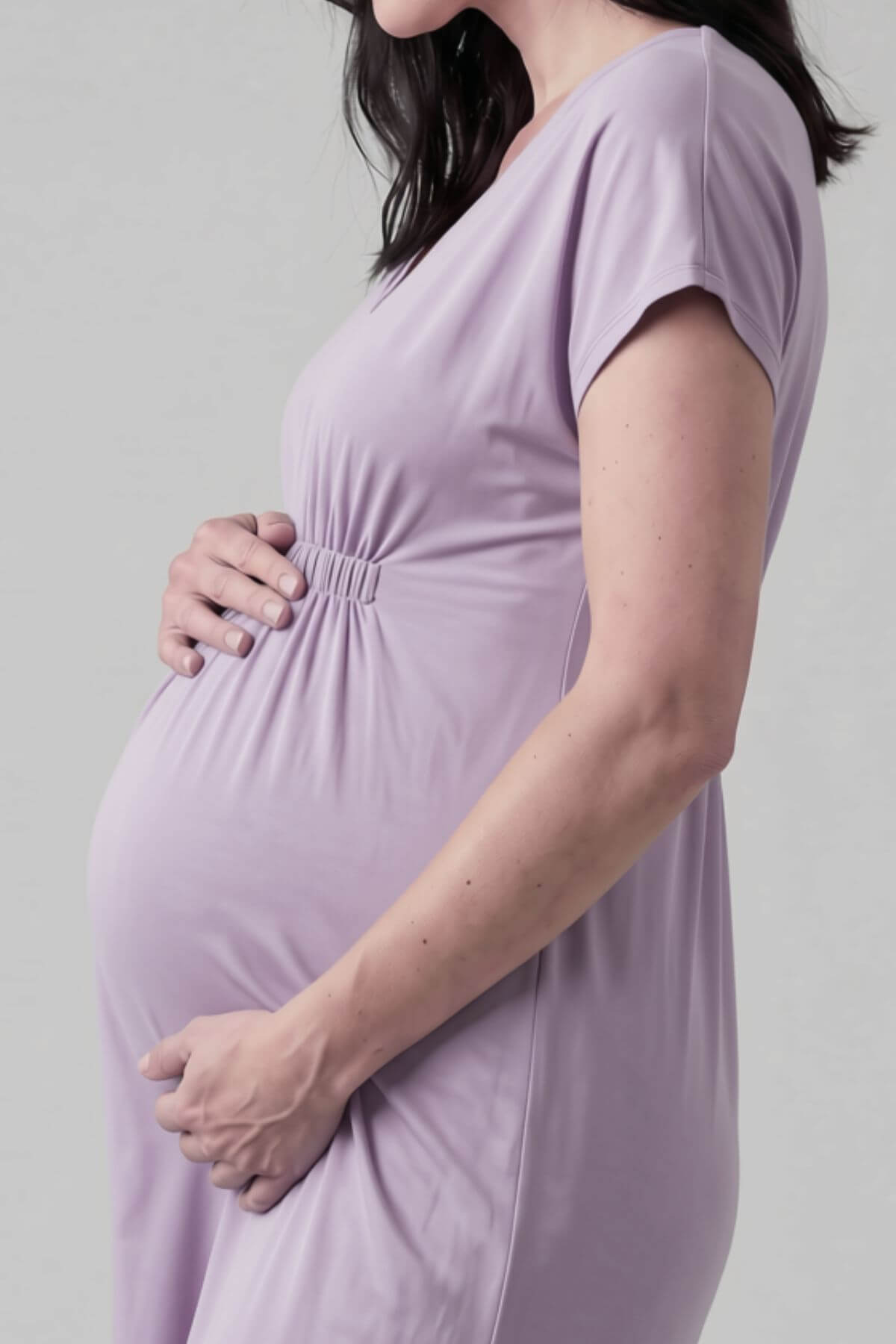 A pregnant woman wearing a light lavender maternity dress, holding her baby bump against a neutral light gray background.