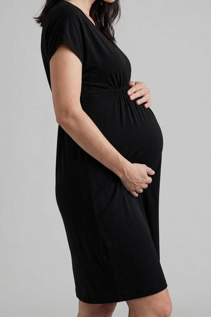 A pregnant woman wearing a black short-sleeve maternity dress with a gathered waist and side pockets, shown in a side-profile view against a solid gray studio background.