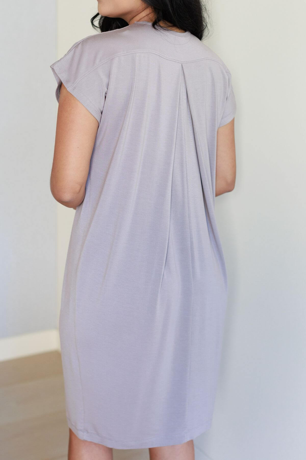 A woman shown from the back wearing a short-sleeve light lavender-gray tunic dress with a gathered back seam, standing against a plain off-white wall.