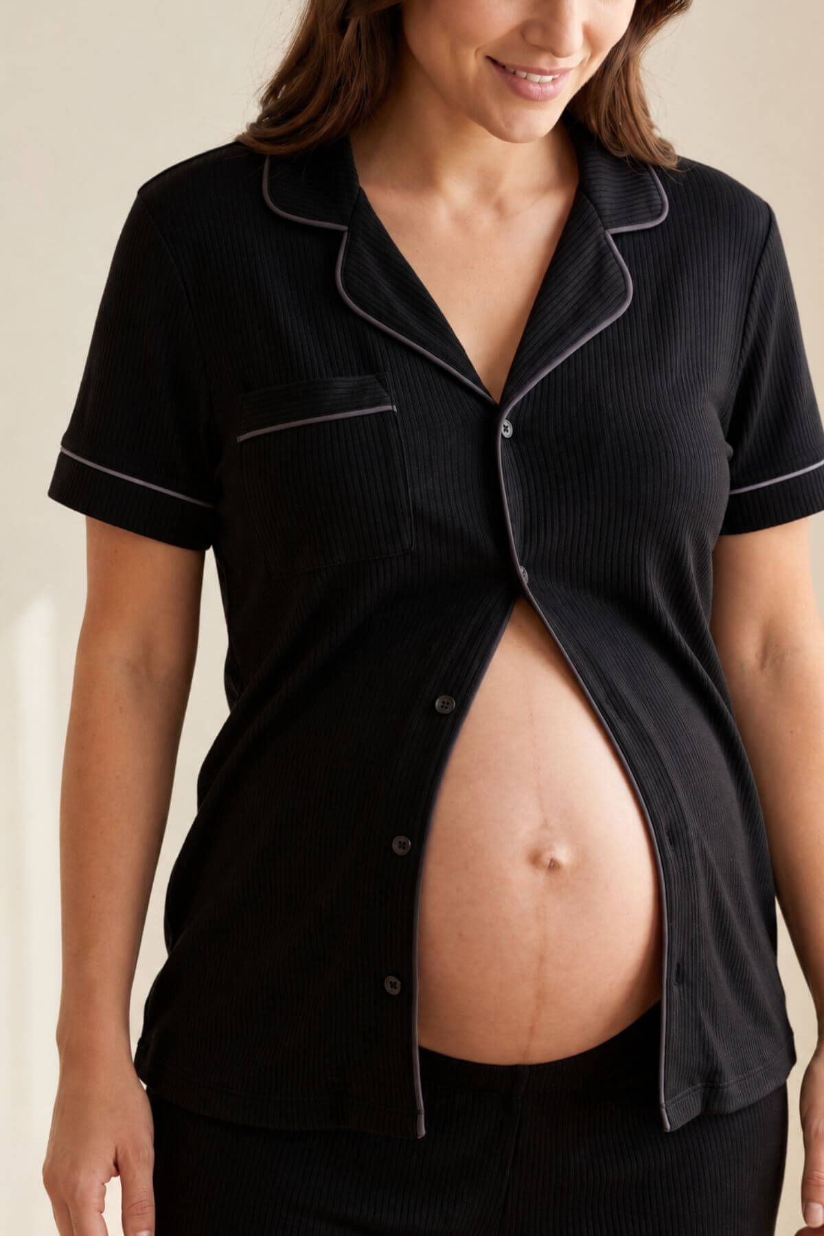 A pregnant woman wearing a black short-sleeve button-front pajama top with light piping trim, shown from the shoulders to the hips against a plain light beige background.s