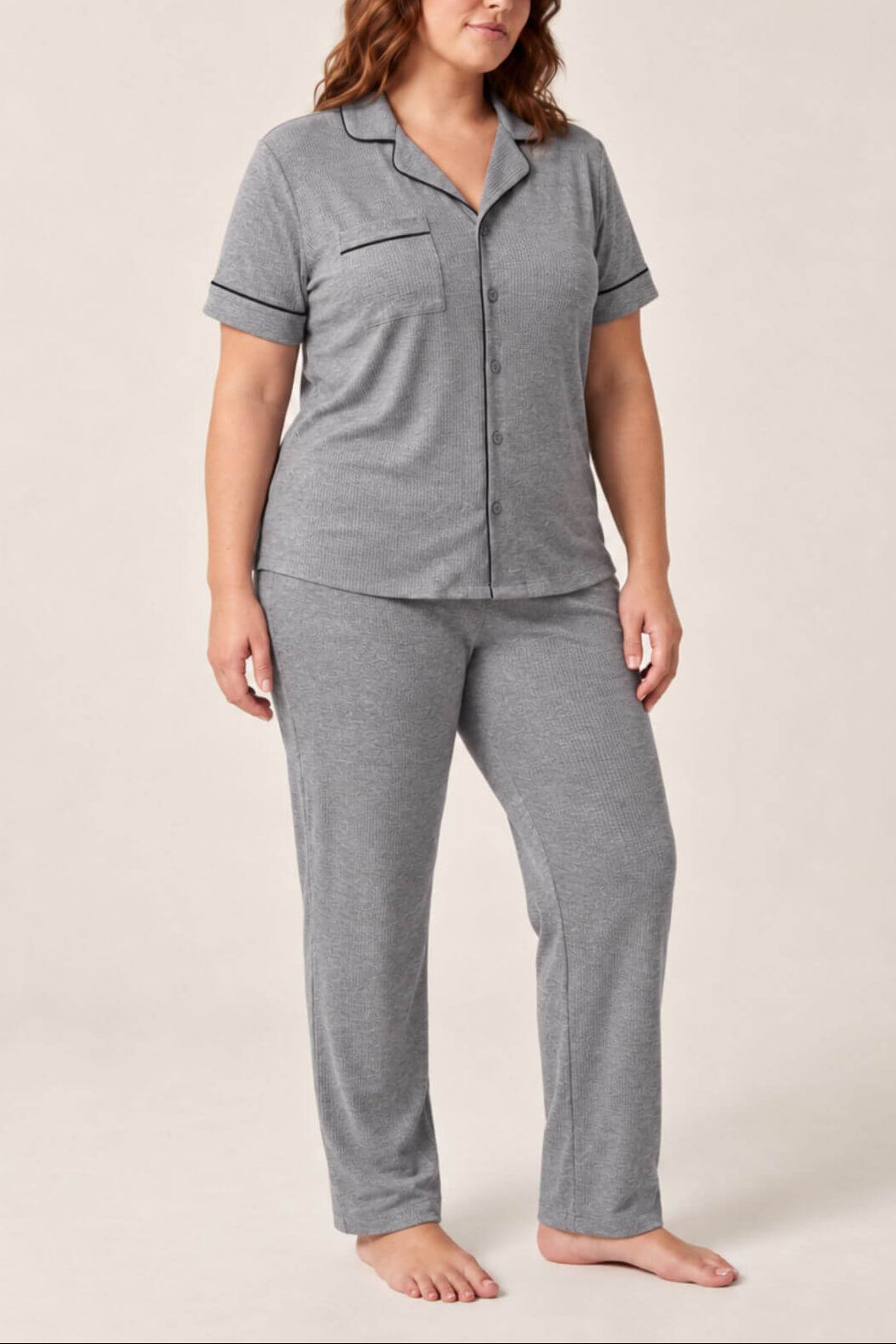 A woman modeling a two-piece heather gray pajama set consisting of a short-sleeve button-front top with a chest pocket and matching long pants, photographed against a plain light beige studio background..