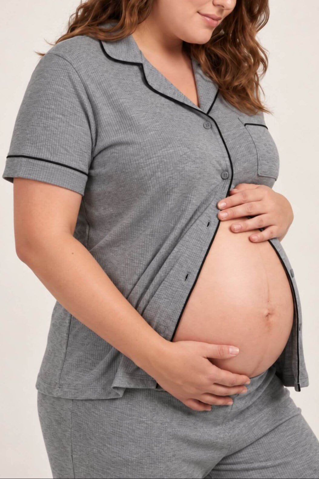 A pregnant woman wearing a gray short-sleeve button-front pajama top with black piping trim, paired with matching gray pants, photographed against a plain light background.
