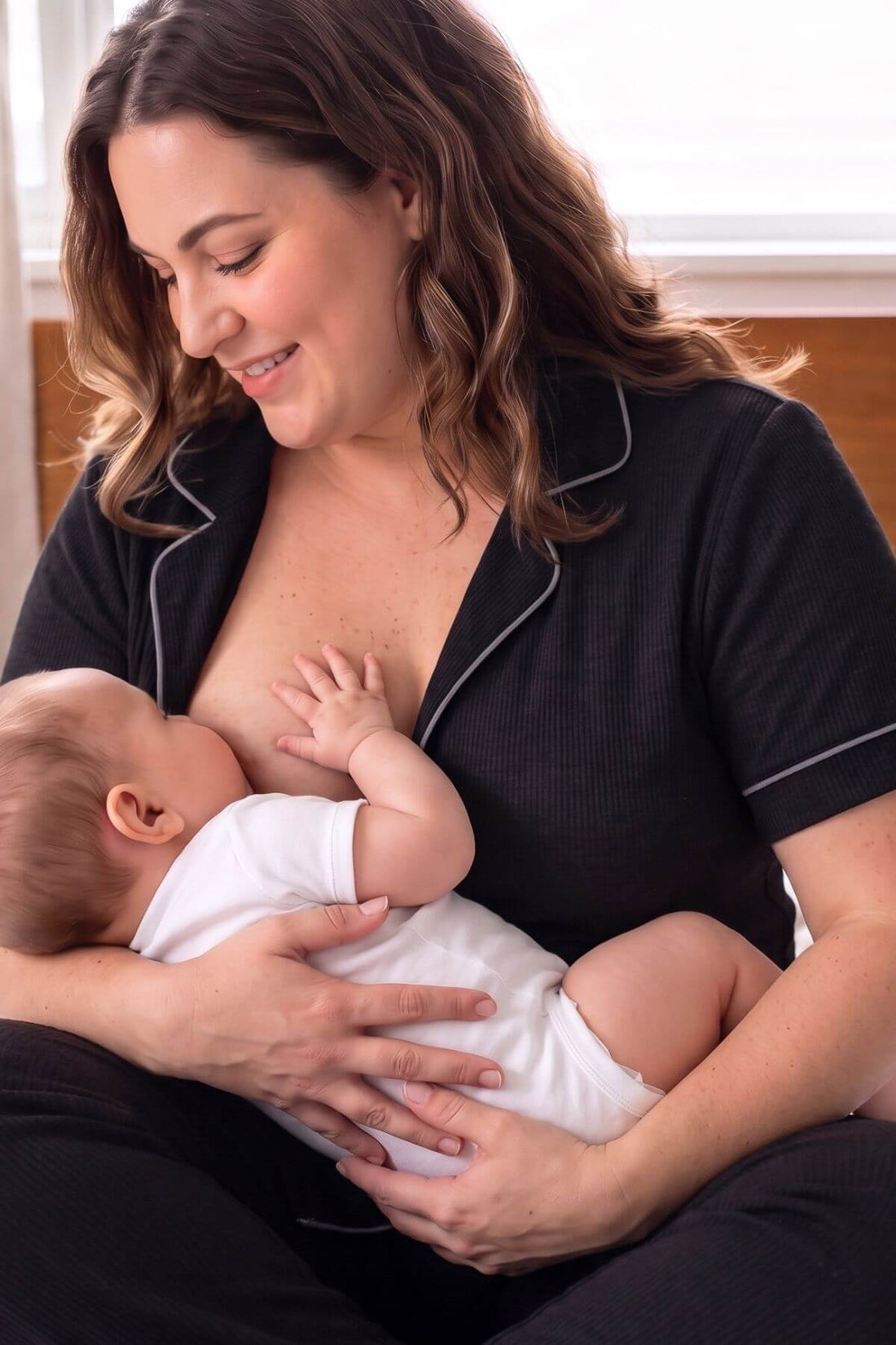 A woman in black pajamas with white piping holds a baby wearing a white onesie while breastfeeding. The scene is shot in a warm, softly lit indoor setting.s