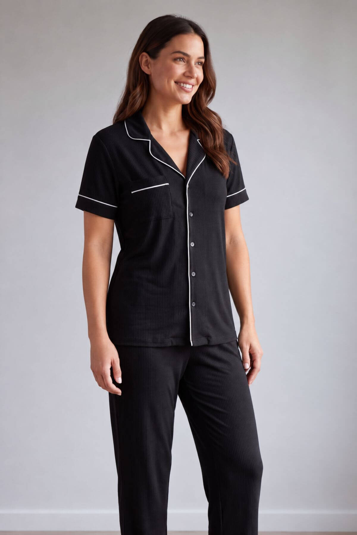A woman modeling a black short-sleeve pajama shirt with white piping trim and matching black pajama pants against a light gray studio background.s