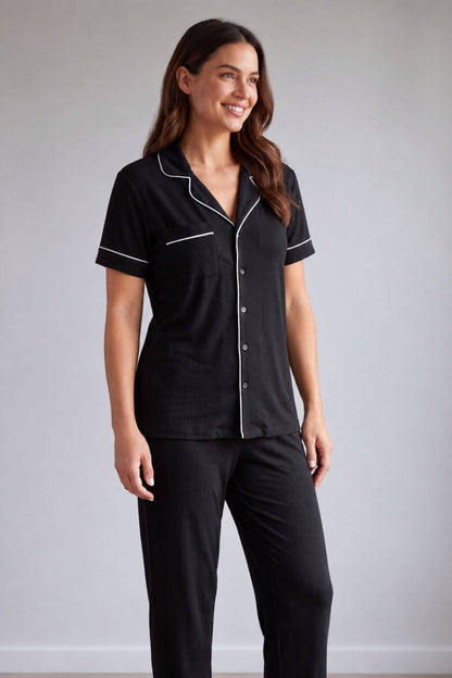 A woman modeling a black short-sleeve pajama shirt with white piping trim and matching black pajama pants against a light gray studio background.s