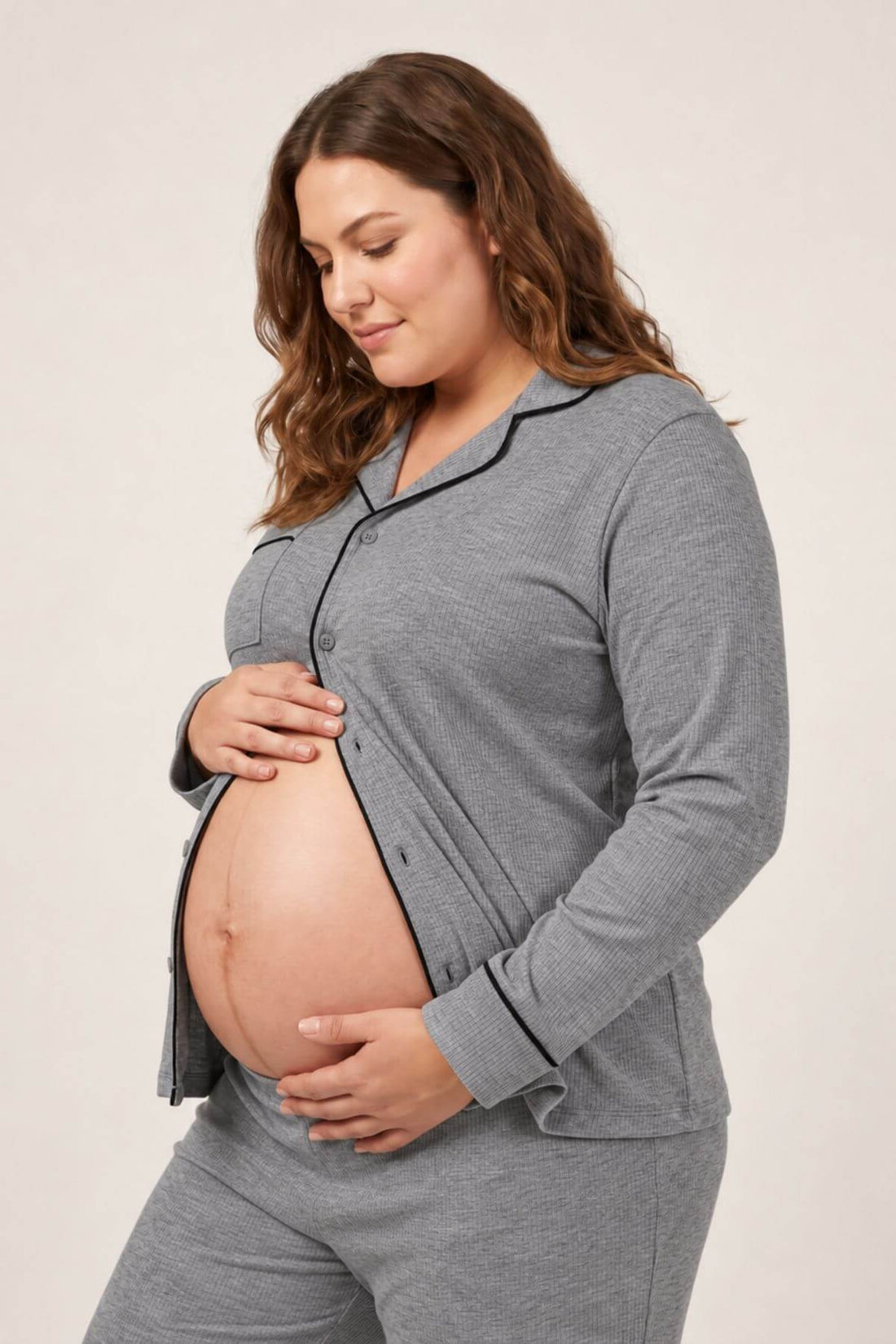 A pregnant woman wearing a heather gray long-sleeve pajama cardigan with black piping and matching gray pajama pants, standing against a light beige studio background.