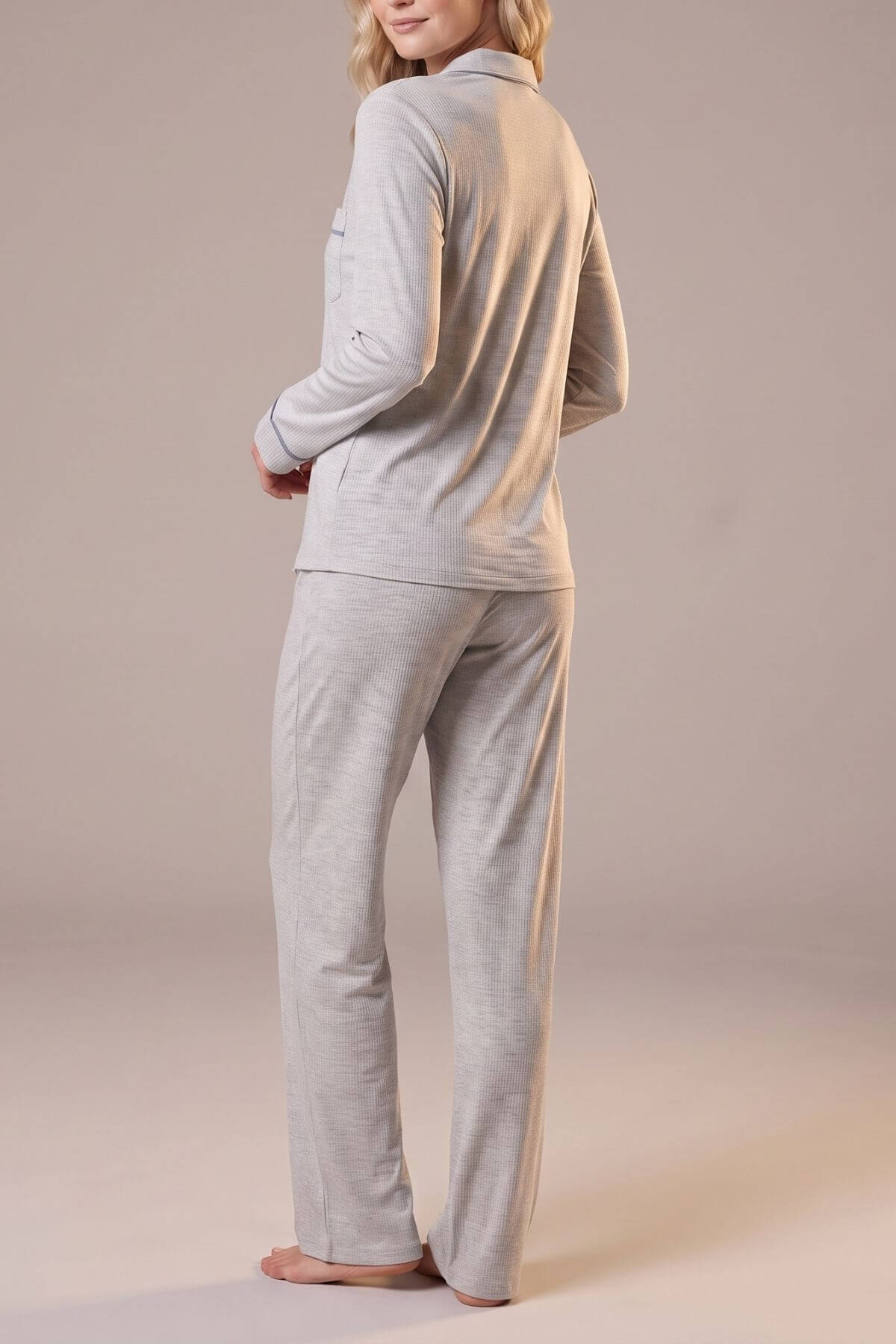 A woman modeling a light gray two-piece pajama set, featuring a long-sleeve collared top and matching straight-leg pants, shown from a rear three-quarter angle against a solid neutral studio backdrop.