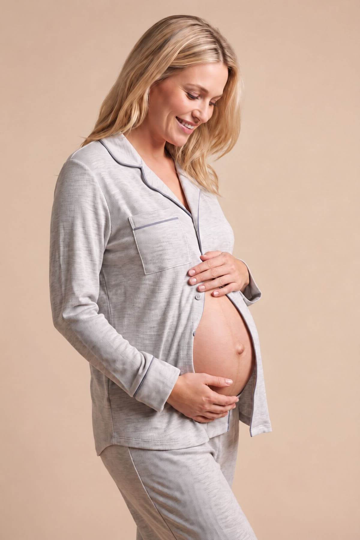 A pregnant woman wearing a light gray long-sleeve pajama set with a zip-up top and matching pants, standing against a solid beige background.
