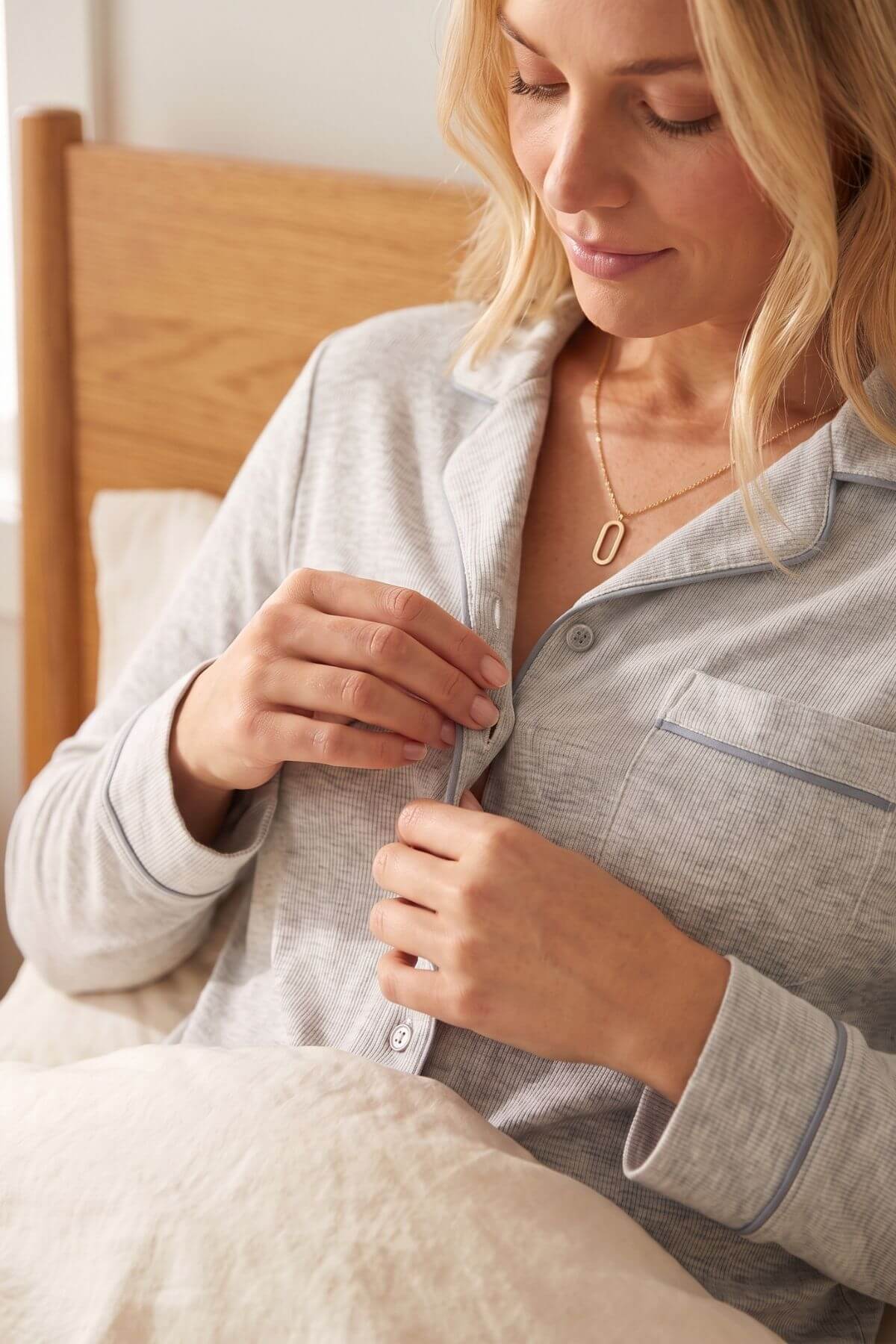 A woman in a bedroom wearing a light gray long-sleeve pajama shirt with thin blue piping, buttoning the collar. She is sitting in bed with a wooden headboard and white bedding visible.