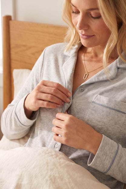 A woman in a bedroom wearing a light gray long-sleeve pajama shirt with thin blue piping, buttoning the collar. She is sitting in bed with a wooden headboard and white bedding visible.