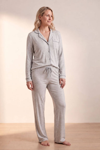 A woman modeling a light gray two-piece pajama set consisting of a long-sleeve button-front top with a chest pocket and matching drawstring pants, standing against a warm beige studio backdrop.