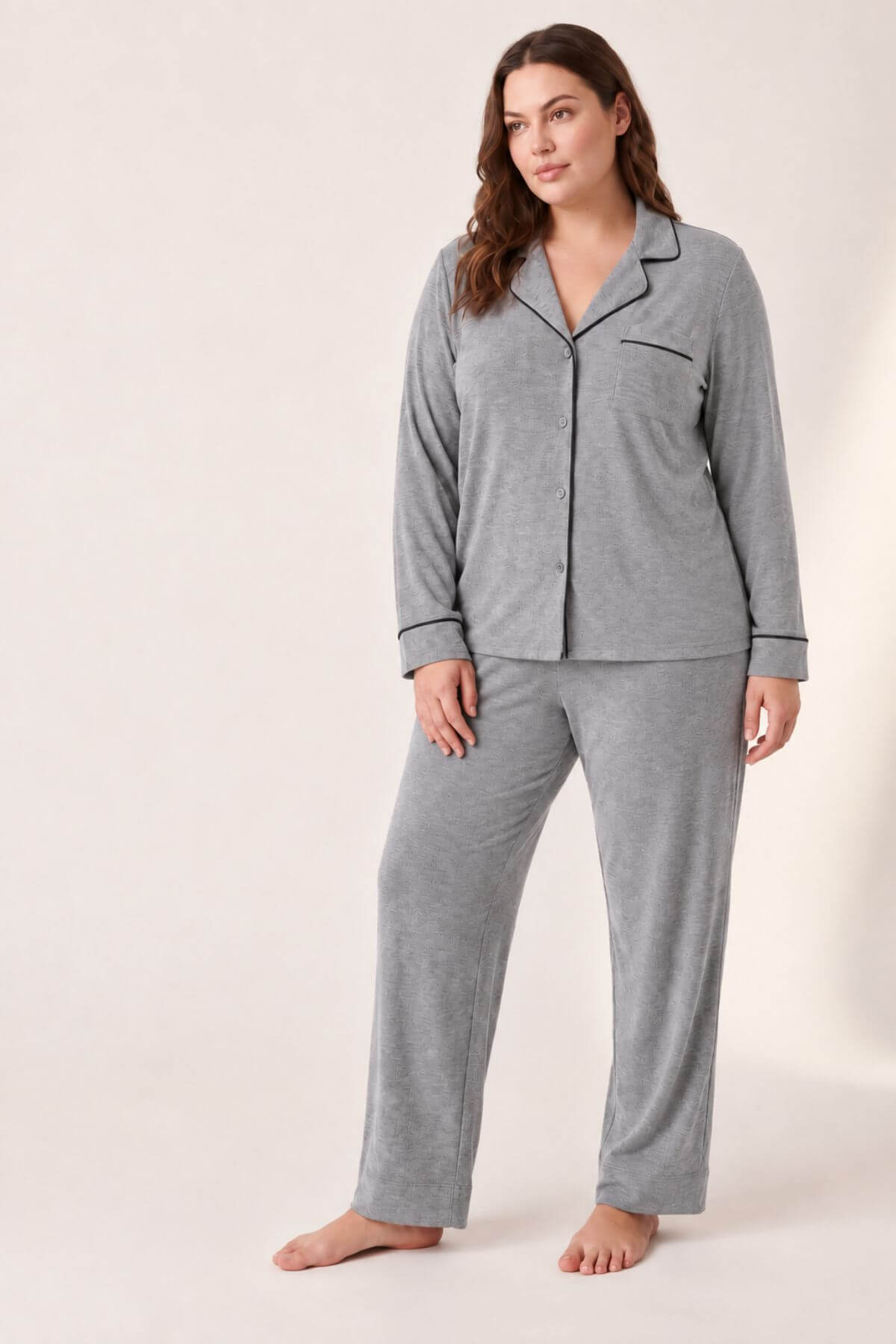 A woman modeling a two-piece gray pajama set with black piping details, consisting of a long-sleeve button-up top and matching pants, against a plain beige studio background.