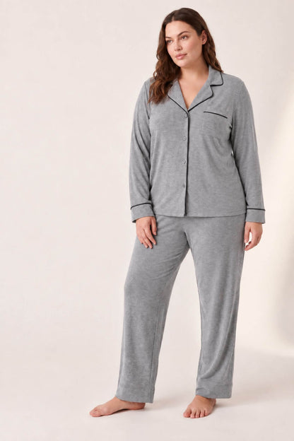 A woman modeling a two-piece gray pajama set with black piping details, consisting of a long-sleeve button-up top and matching pants, against a plain beige studio background.