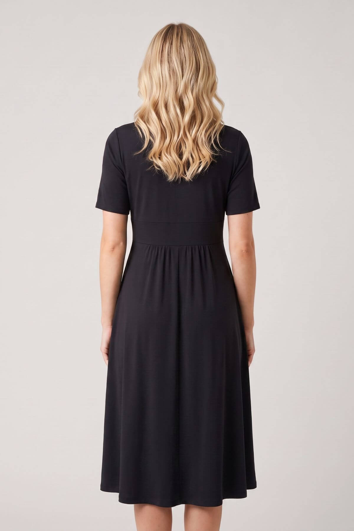 A woman viewed from the back wearing a short-sleeve black midi dress against a plain light gray studio background.