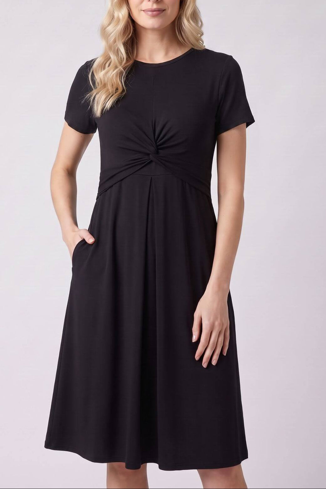 A woman modeling a black short-sleeve A-line dress with a gathered twist detail at the waist and side pockets, photographed against a light gray studio background..