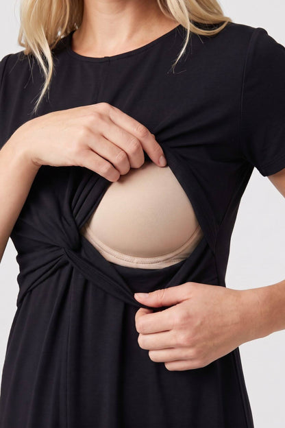 A close-up of a woman wearing a black short-sleeve maternity dress, lifting the fabric to reveal a beige maternity bra underneath, set against a plain light gray background..
