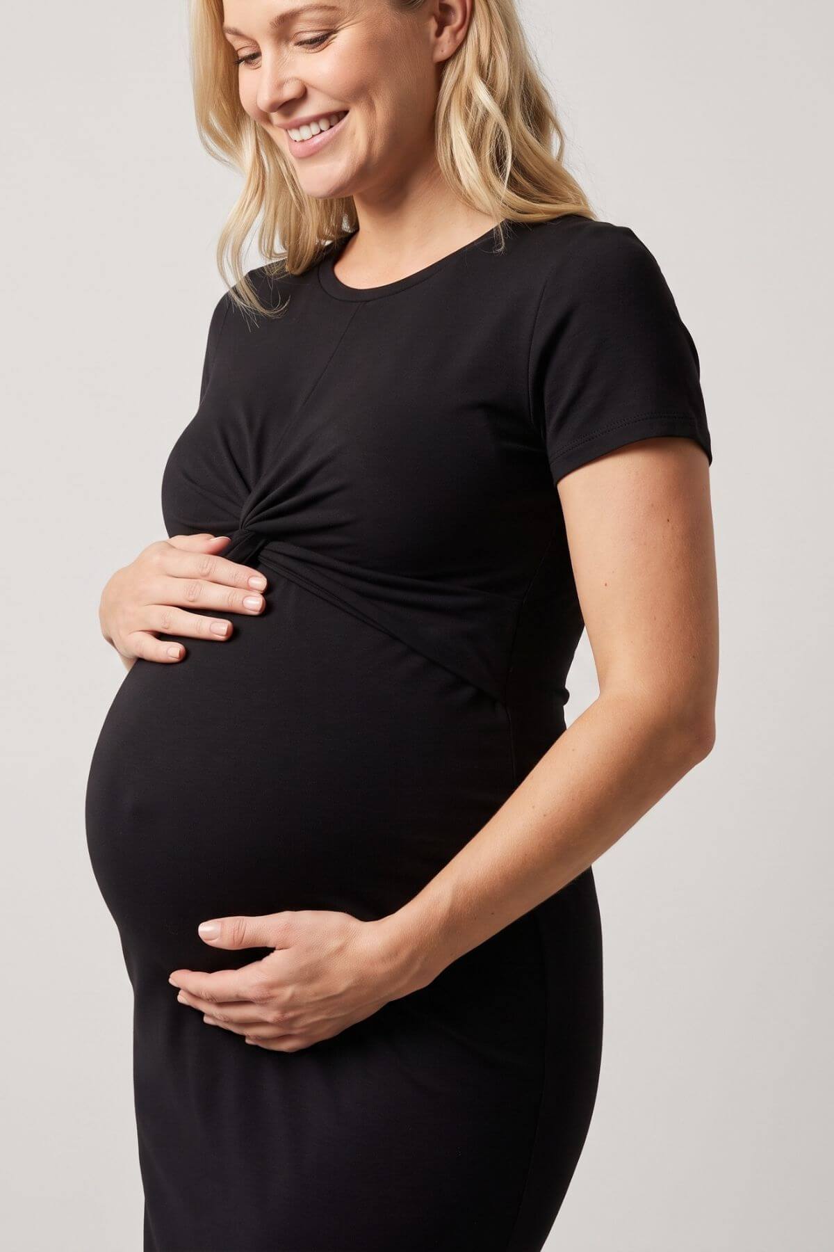 A pregnant woman wearing a black short-sleeve maternity dress with a twisted front detail, posing with her hands on her belly against a light gray background..