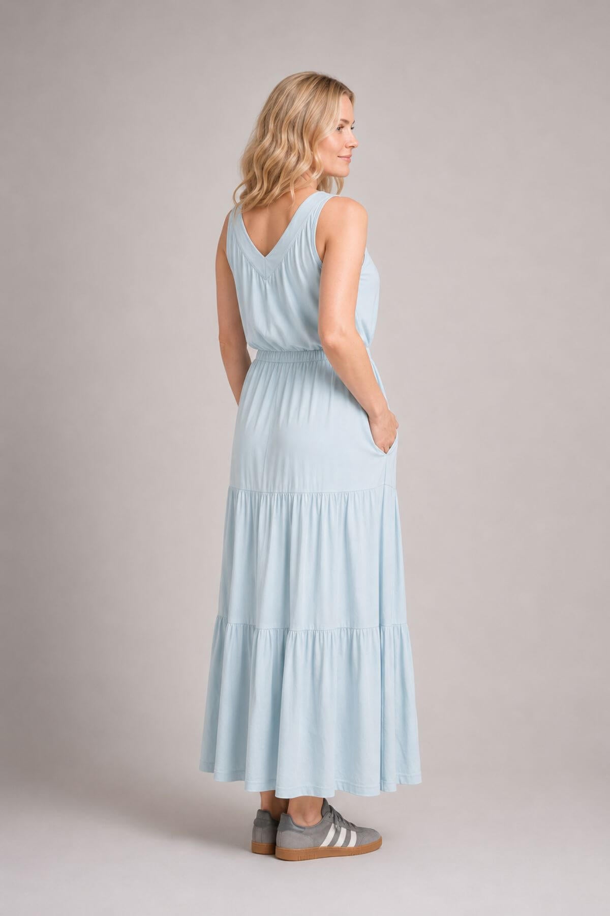 A woman modeling a light blue sleeveless maxi dress from the back, standing against a neutral gray studio background.