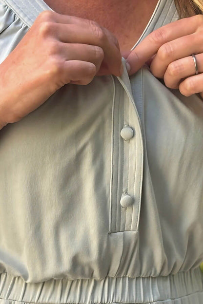 Close-up of a person wearing a light sage green or gray-green top, pulling the front placket open with both hands. The top has a gathered waist and a vertical button placket with two light-colored buttons.