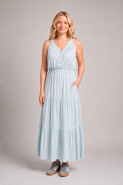 A woman modeling a sleeveless light blue tiered maxi dress with a V-neckline and button detail at the bust, standing against a neutral gray studio background.