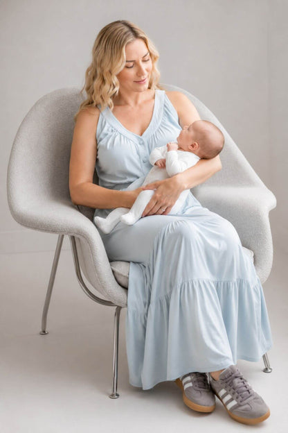 A woman in a light blue sleeveless maxi dress sits in a modern gray chair, holding a baby dressed in white. She is wearing gray sneakers with white stripes.