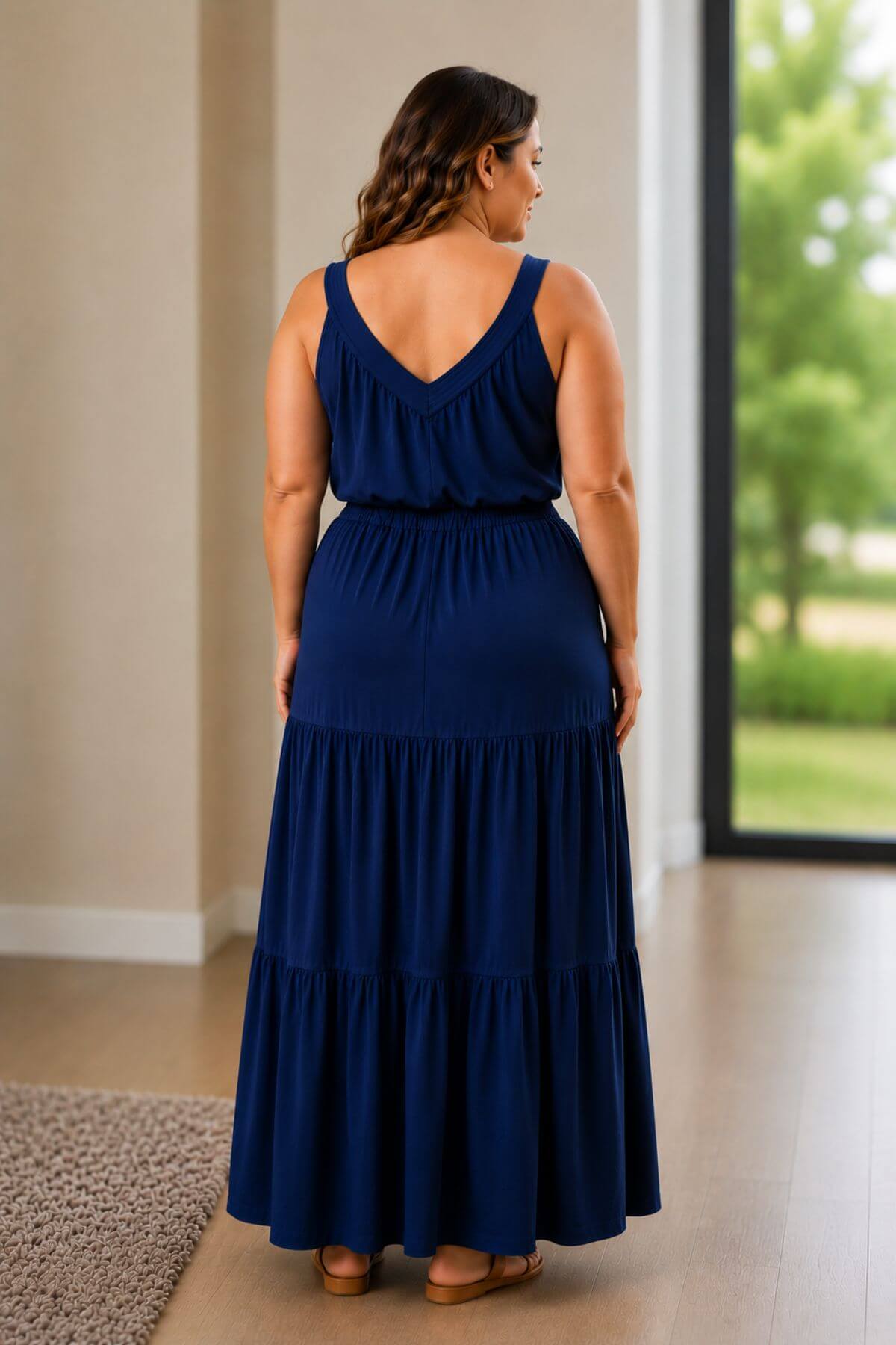 A woman wearing a navy blue tiered maxi dress, shown from the back in a bright indoor setting with a large window showing green trees outside.