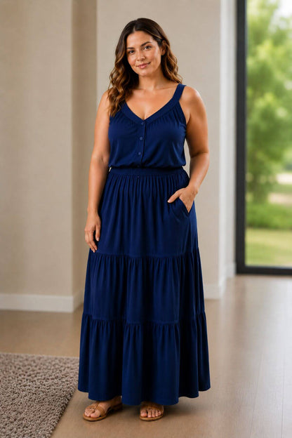 A woman modeling a sleeveless navy blue maxi dress with a tiered skirt and buttoned V-neck bodice, standing in a bright indoor room with a window view of greenery.