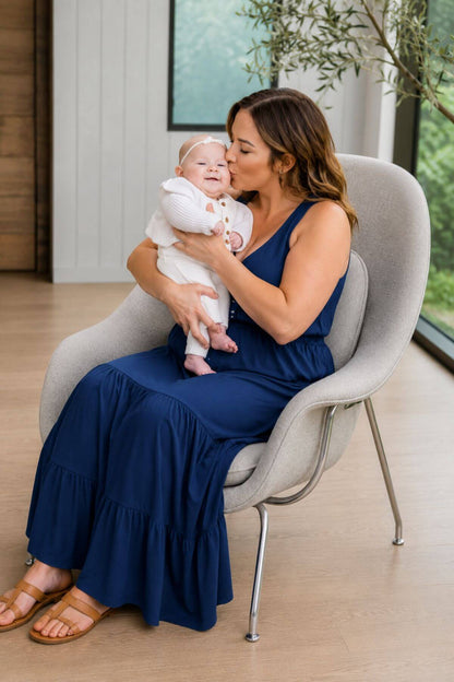 A woman in a navy blue sleeveless maxi dress sits in a light gray modern chair, holding and kissing a baby dressed in white. The setting is a bright, modern room with light wood floors and large windows.