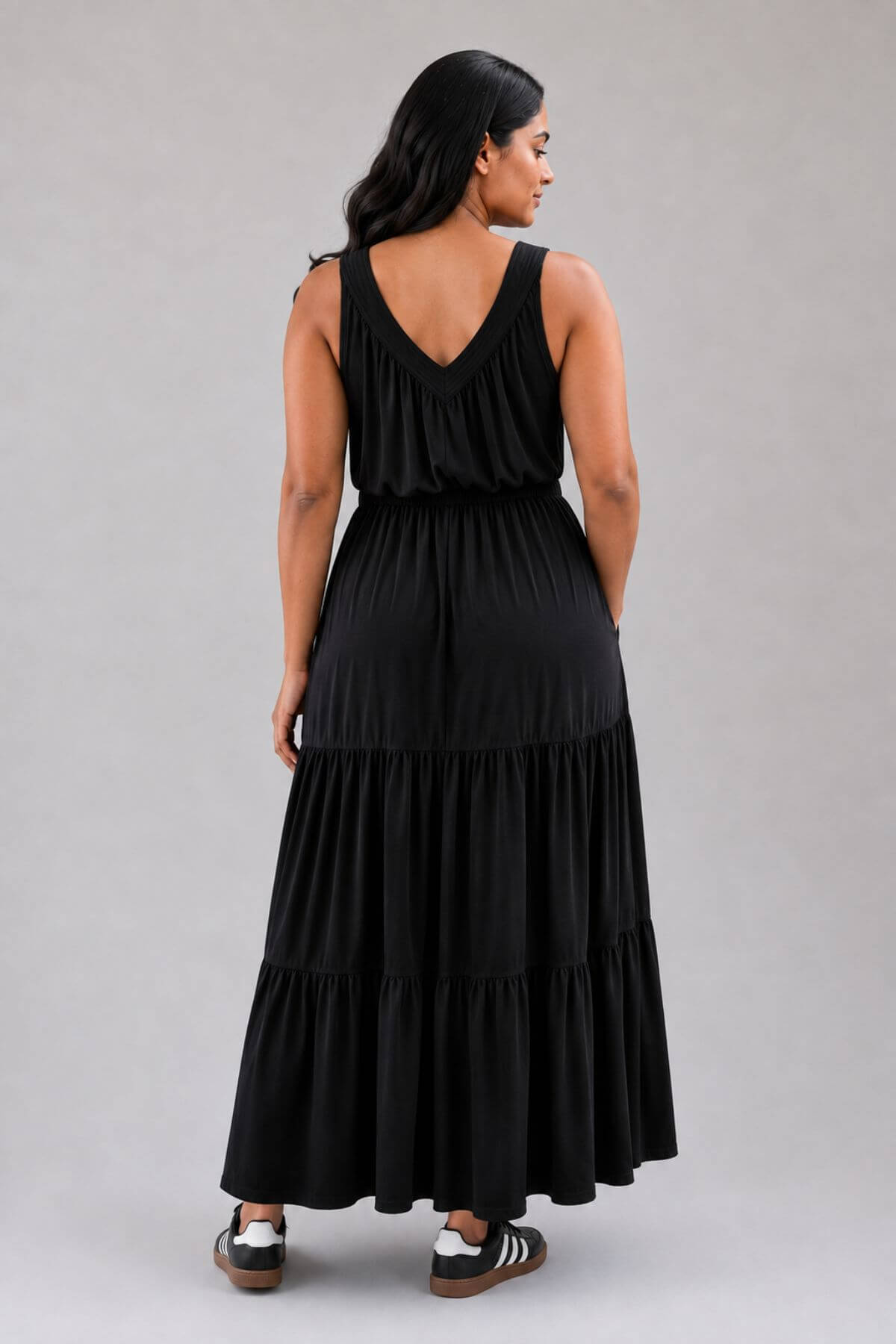 A woman wearing a black sleeveless tiered maxi dress, shown from the back against a solid gray studio background.