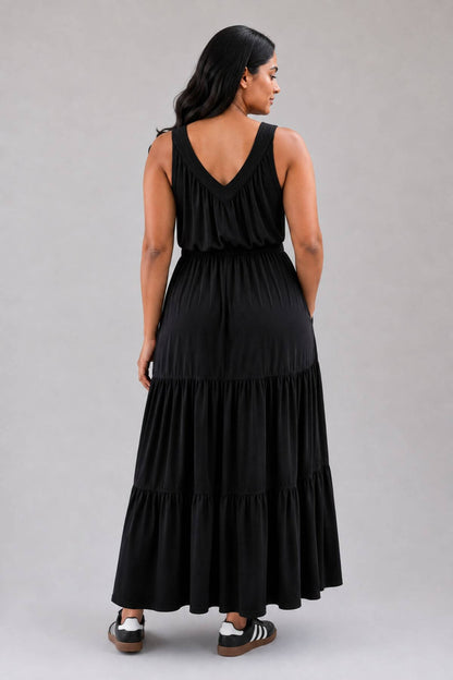 A woman wearing a black sleeveless tiered maxi dress, shown from the back against a solid gray studio background.