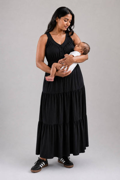 A woman wearing a black sleeveless tiered maxi dress holds a baby dressed in a white outfit. She is standing against a solid gray studio background.