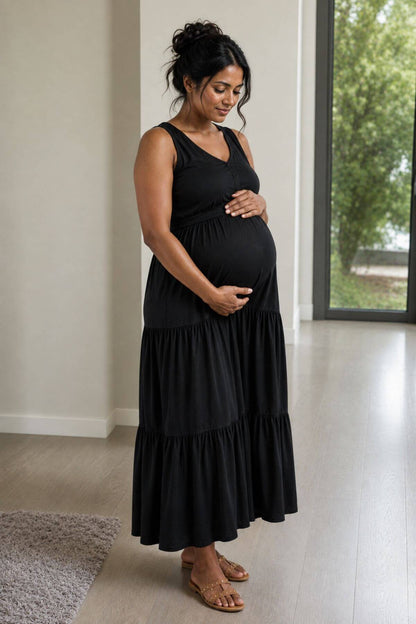 A pregnant woman wearing a black sleeveless tiered maxi dress and brown sandals, standing in a bright room with light wood floors and a window.