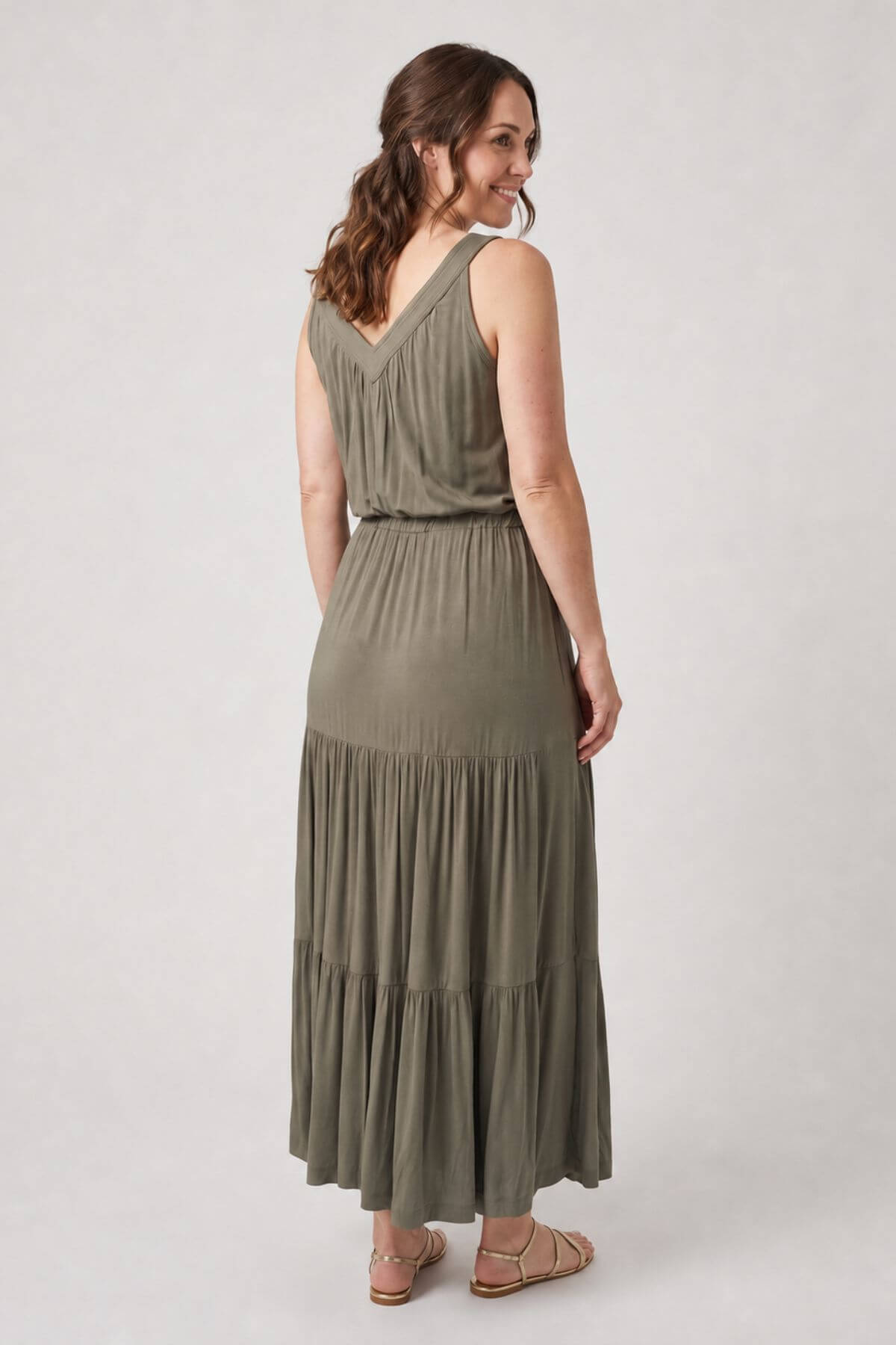 A woman modeling a sleeveless olive green maxi dress with a tiered skirt, shown from the back against a light gray studio background.