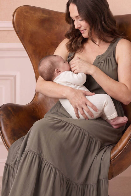 A woman in a sleeveless olive green dress sits in a brown leather armchair, breastfeeding a baby wearing a white onesie.