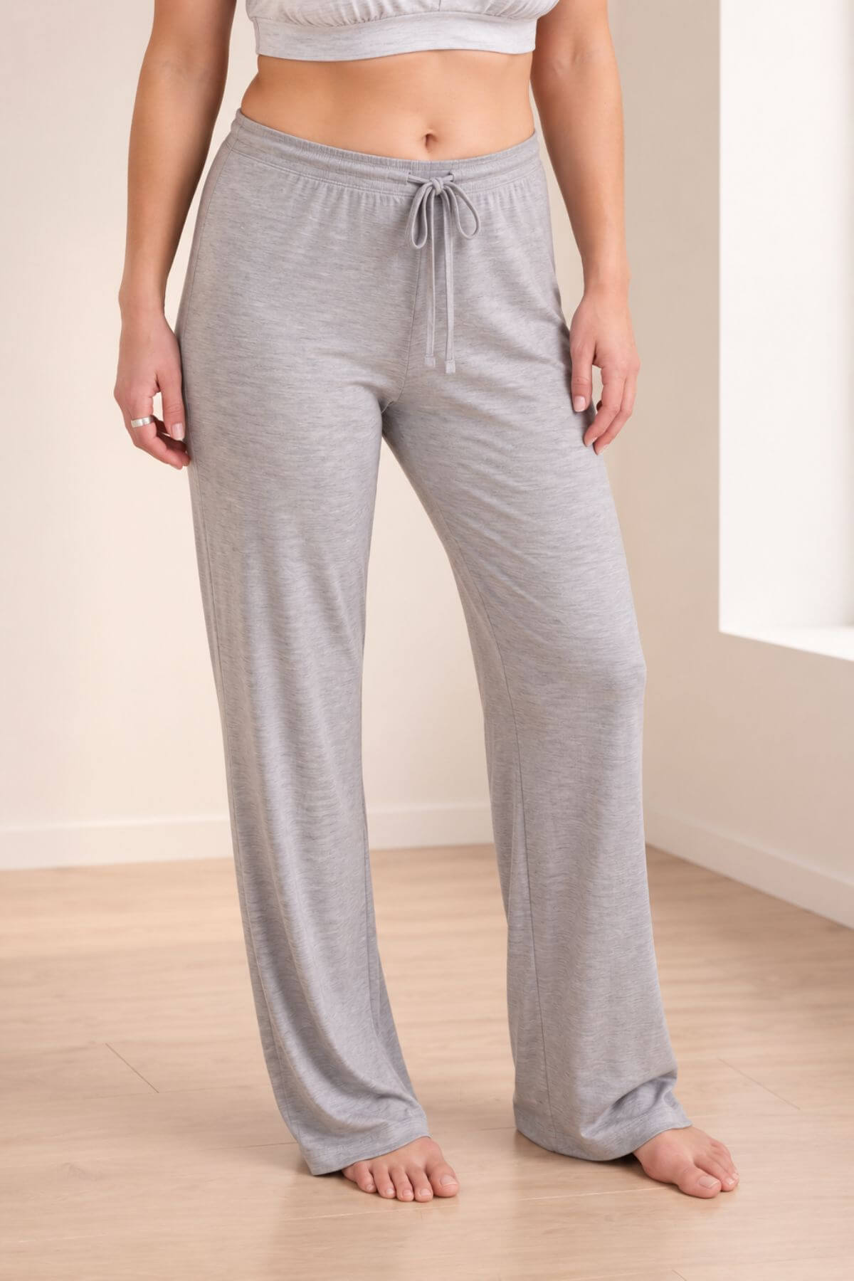 A person wearing light heather gray drawstring lounge pants, shown from the waist down in a bright indoor room with light wood flooring.